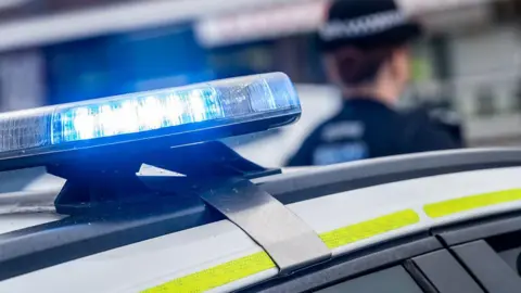 A stock images of the top of a white police car with yellow reflective strips on. The blue emergency lights are turned on. In the background is a female police officer with her back turned away from the camera. She is wearing a black police uniform including black police hat.