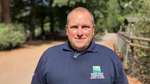 A man in a blue sweatshirt stands in front of a wooded area at the River Dart Country Park in Devon