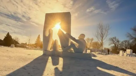 Justin Scott A snow sculpture in a snowy field depicts a wall with a 2D cut out of a person. Next to it is a 3D person holding their hand up to the 2D cut down. Sun is shining through the 2D cut out. Other sculptures can be seen int he distance as well as winter trees.