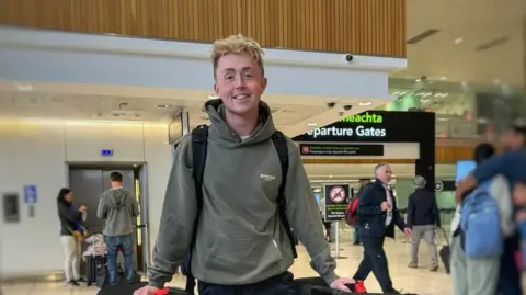 Jamie Online Vintage Jamie Harkin is standing in the airport departures area. He is wearing a green hoodie and wearing a black bag pack. There is an elevator behind him with two people standing outside of it. To his side there are four people with two hugging. In the background there is a large black departures gate.  