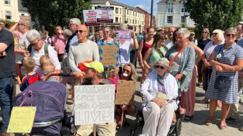 A large group of people, including some with prams, one sitting in a wheelchair and some children. About half of them are holding banners and placards - one reads Maternity cuts cost mums and babies lives, another reads Save north Devon's maternity services, and another reads Maternity cuts cost lives.