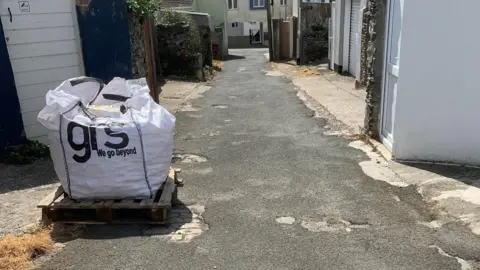 Marie Swords A pallet of stones in a big white bag. It's on a concrete back street with buildings and garages either side.
