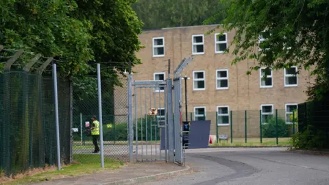 RAF Linton-On-Ouse in North Yorkshire. A metal gate with barbed‑wire fencing stands partly open, with a person in a high‑visibility vest inside the compound and a brick building surrounded by trees in the background.