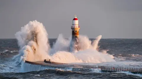 Getty Images Roker Pier. Large waves are crashing into the pier and it's lighthouse. The pier curves to the right and has railings along each side. The lighthouse has a white and red dome.