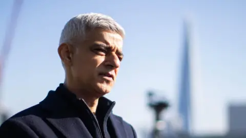 Sadiq Khan, a man with short grey hair wearing a blue coat and zip jacket. He looks like he is speaking and there is a blurry city skyline behind him. The sun is on his face.