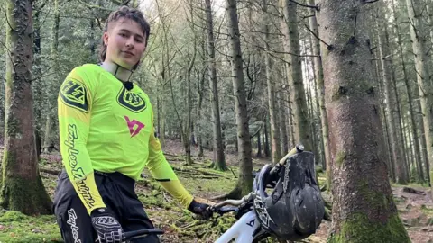 A man is stood in a forest next to a mountain bike. He is wearing a bright yellow jersey, black trousers and black and white gloves. He has brown hair in a mullet style. He is looking at the camera with a slight smile. 