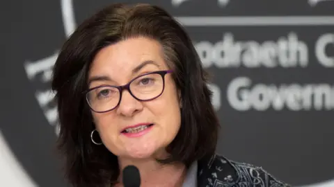 Eluned Morgan speaking at a Welsh government press conference, she is looking at the camera and wearing purple glasses and pink lipstick