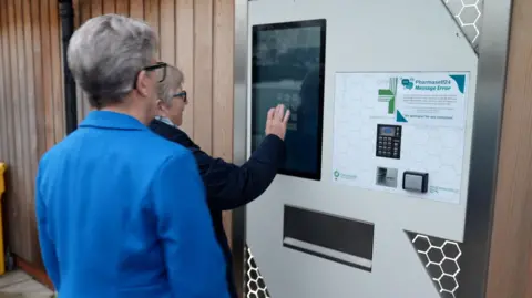 Two people stand outside the wooden walls of a building at an automated prescription collection machine. It resembles a vending machine with a white front, a large computer screen, a white board with keypad and instructions and a metal letterbox. A woman is using the touchscreen to operate the system. She has short grey hair, and dark-rimmed glasses and is wearing a dark blue jacket. A second woman looks on. She is wearing a vivid blue jacket and has short grey hair and glasses.