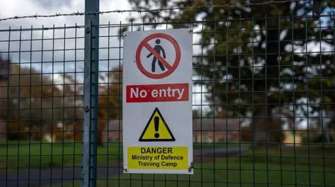 Getty Images A warning sign reading "No entry" and "Danger Ministry of Defence Training Camp" is attached to a wire fence topped with barbed wire. Open grassland can be seen beyond the fence.
