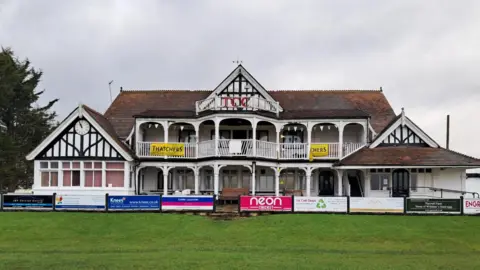 A two-storey building in a mock Elizabethan style, with balconies and in front a fence with sponsors for the cricket club.