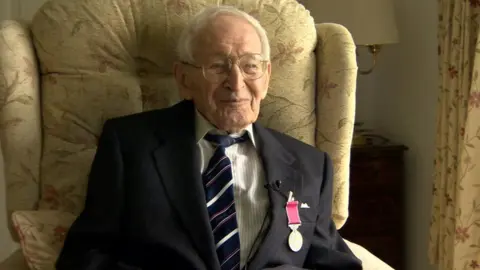 100 Peter Davis sitting in an armchair. He has white hair and big glasses and a smile. He wears a blue blazer over a white shirt and striped tie. On his left lapel is the British Empire medal.