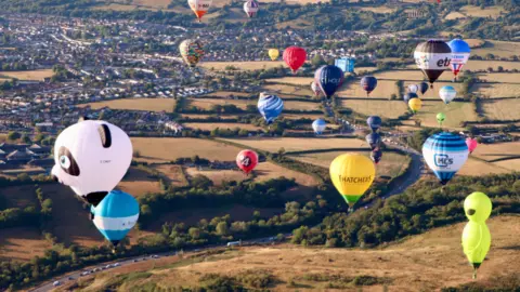 Many colourful balloons are seen flying over the outskirts Bristol and yellow/brown fields