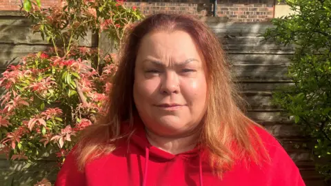A woman with red-coloured shoulder length hair and wearing a red hoodie stands outside in a garden in front of a wooden fence. 