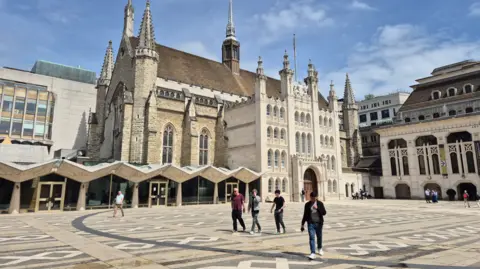 A large historic stone church with Gothic architecture stands beside an open paved courtyard under a bright blue sky with scattered clouds. The building features tall spires, pointed arched windows, and a pale decorative facade.