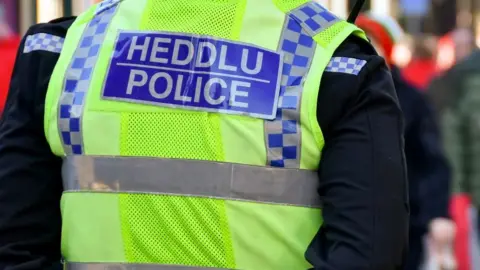 The back of a police officer wearing black and yellow high-vis uniform which reads: Heddlu - Police. 