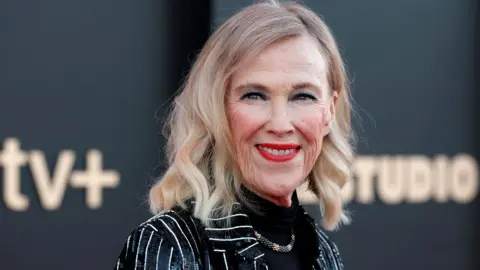 Catherine O'Hara, who has a blonde bob and wears red lipstick, a black poloneck top and a sparkly pinstriped blazer, photographed smiling on a red carpet in 2025