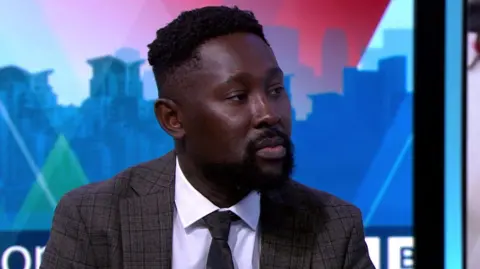Jason Lartey seen in a suit and tie sitting in a television studio, looking to his right, with a blurred cityscape graphic in the background.