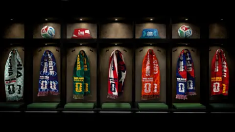 Hand Drawn Pixels A picture of several scarves in various colours lined up in what looks like a football team's dressing room. There are footballs and bucket hats above them. 