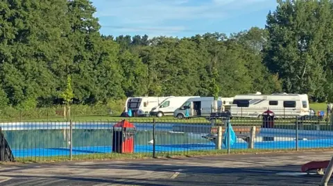 A large outdoor paddling pool in a children's park. There are several camper vans and cars on the grass to the side.