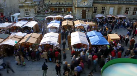 B&NES Council About a dozen wooden chalets some with plastic coverings on the roof, and lots of people around the stalls outside the Romam Baths