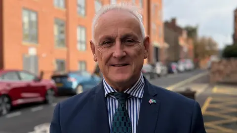 BBC Alan Graves on a city street wearing a blue suit jacket, blue striped shirt and blue & green tie. His lapel pin features Union and Derbyshire flags crossed.