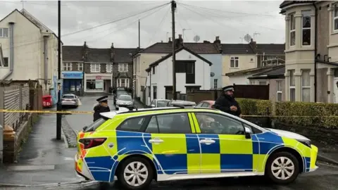 Bishop Road blocked by a police cordon. A police car is in the foreground of the picture. There are two police officers in uniform next to the car. Yellow cordon tape is also across the street at both ends. It is a grey day and shops  at the end of the street are closed. 