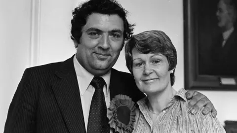 Pacemaker John and Pat Hume are pictured together. He is on the right, wearing a suit and large rosette. Pat, on the left is smiling. His arm is on her shoulder.