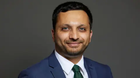 House of Commons/Laurie Noble Professional studio headshot of a person wearing a blue suit jacket, white shirt, and green tie, facing the camera against a dark gray background. The portrait is sharply focused with even lighting and a neutral backdrop.