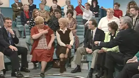 Haney is in the audience of the TV show Kilroy. A man with a gray -haired man in a suit sits to the right of the image. Haney seems to be trying to intervene while bringing a woman to the left. All spectators are looking at Haney. He wears a red skirt and jacket and points to the woman next to him.