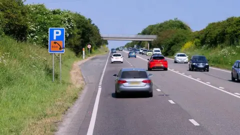 Cars travel on both sides of the A30 near Connor Downs in Cornwall on a sunny day. A layby with a sign for parking and SOS phone is on the left-hand side. A bridge crossing over the road is in the distance.
