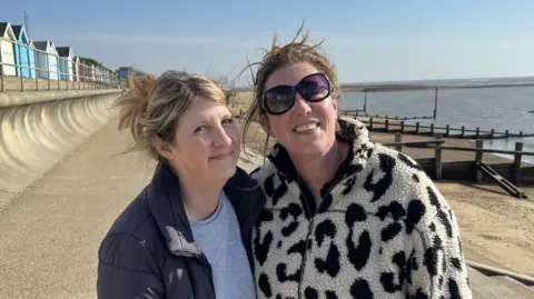 George King/BBC Imogen Ainsley and Jo Elliot standing side-by-side on the Felixstowe seafront. The beach and sea can be seen to the right, while beach huts can be seen in the background to the left.