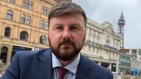 Chris Webb MP with brown hair and beard wearing a navy blue suit and red tie standing outside Blackpool Winter Gardens with Blackpool Tower in the background.