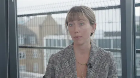 Antonia Jennings wearing a checked blazer and necklace, seated indoors in front of a large window with metal railings visible outside, speaking during an interview.