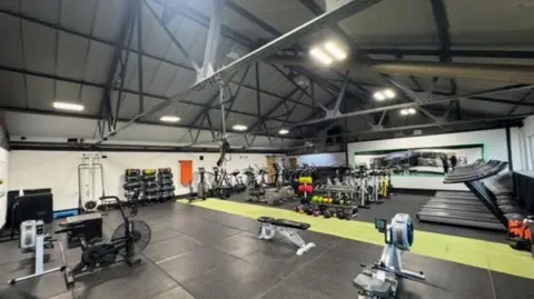Chloe Cartwright A gym with a triangular ceiling has metal beams running across the width of the space. On the floor is a selection of gym equipment including rowing machines, treadmills and a weight section with benches.