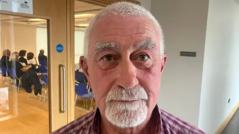 A man with short grey hair, grey eyebrows and grey facial hairs stares at the camera as he stands in front of a door. He is wearing a purple checked shirt.
