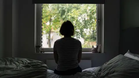 Getty Images The silhouette of a woman sitting on her bed looking out her bedroom window at trees. She is wearing a white top with thin black stripes.