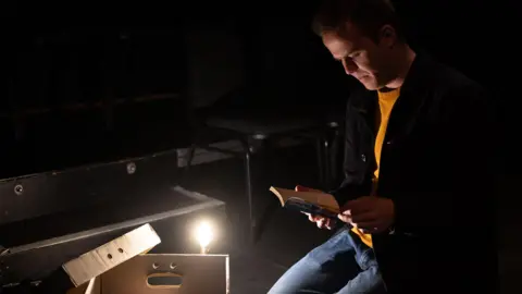 Frederick Houlahan A man kneeling on a theatre stage with dark lighting while reading a book