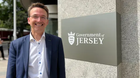 A man with brown hair styled in a quiff smiling at the camera. He has glasses on. He is wearing a dark blue blazer and a white shirt, which has its top button undone. The shirt buttons are blue. He is standing next to a sign that says 'Government of Jersey'.