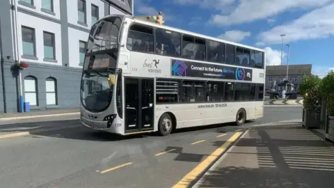A silver double-decker bus drives on a road next to a roundabout and large building.