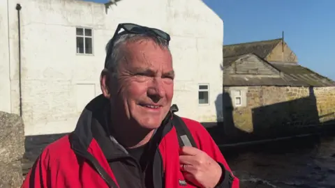 John Brooks- a older man with short grey hair, sunglasses on the top of his head and wearing a red jacket - smiles in the sunshine.