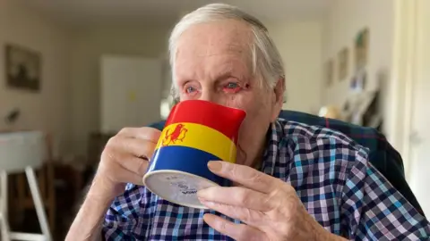 Leslie Lemon, with a blue, yellow and red custard jug featuring three birds, held to his lips. He is wearing a blue checked shirt and has short grey hair. 