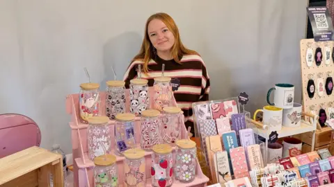 Alaina Davis smiles at the camera as she stands behind a stall that is displaying her products, such as colourful water bottles, mugs and keychains. She has auburn hair and is wearing a striped jumper. 