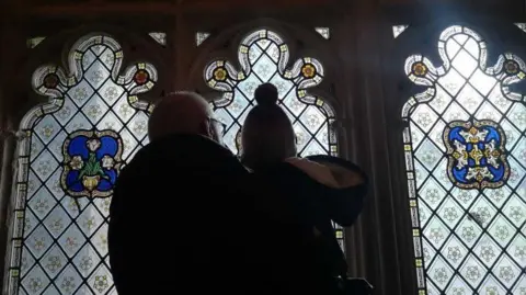 PA Media Mr Berry and one of his grandchildren pictured from behind, looking up at a stained glass window in a church. They are in silhouette and there is bright light shining through the three ornate panels of glass. 