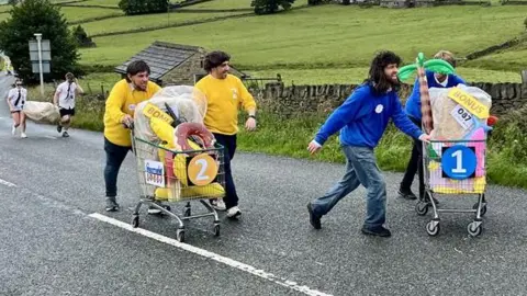 BBC/Dave Edwards Four participants in wigs push two shopping carts loaded with various items uphill. On the right, two wear blue jumpers, while those on the left sport yellow. In the background, two other runners—dressed in sunglasses, white shirts, black ties, and shorts—can be seen carrying a bale of straw between them as they tackle the hill.