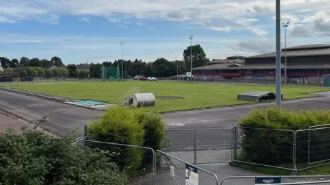 A view over security fencing towards Queens Park and its excavated running track.
