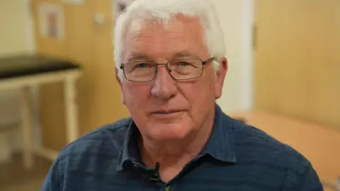 Ged Cashley is wearing a blue shirt and glasses. The 72 year old suffered a cardiac arrest in 2018 and now has annual health checks.