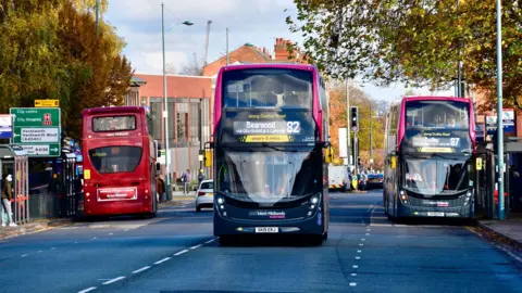 Two black and pink buses are travelling on a three lane road towards the camera. There is a red bus at a bus stop on the left travelling in the opposite direction