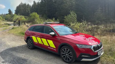 A red fire car with yellow stripes on the side is parked on the side of a gravel path on a grass verge. In the background the path continues, around an area of woodland with numerous trees. It is a sunny day.