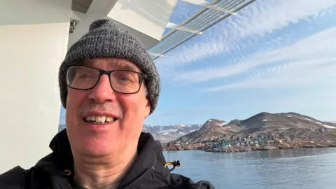 Mike is wearing a grey wooly hat and a black waterproof jacket. Over his left shoulder is the village of Ittoqqortoormiit.
