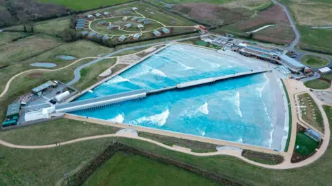 The Wave, taken from an aerial view high up in the sky. The facility is a large triangular shape, with one curved edge, bisected by what looks like a pier. There are five even waves running down each edge of the lake. The main facility is surrounded by curved walkways and a number of different buildings, as well as green fields. 